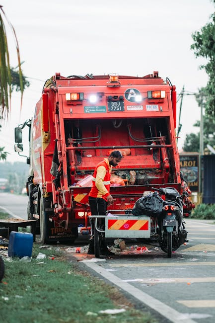 A sanitation worker is seen from the side, wearing a yellow high-visibility vest with red accents, as they operate a large red rubbish collection truck parked on the side of a paved street during daytime. The back of the truck features mechanical components and a clear windowed compartment, with reflective yellow and red stripes on the lower part for visibility. The worker is handling a black plastic bag filled with waste, which is placed on a small metal platform attached to a scooter parked adjacent to the truck. In the background, timber utility poles and overhead power lines are visible, along with some trees and a blue building. The scene captures a moment of on-site waste collection, with debris and waste materials evident on the ground near the truck, emphasizing the context of rubbish removal services typical of independent waste collection or private disposal operations. The overcast sky contributes to diffuse natural lighting, highlighting the textures of the waste and the metallic surfaces of the truck, with some grass and street markings framing the scene, consistent with professional rubbish removal activities in urban or suburban environments. House Clearance Streatham specializes in rubbish and waste management, evident through the visible equipment and operational setup.
