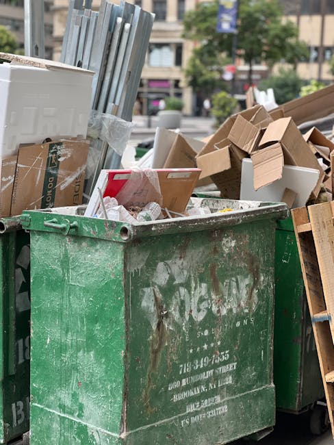 A large, open-top green waste container with visible signs of dirt and rust, filled with various discarded items including cardboard boxes, white foam packaging, and small plastic containers. The cardboard boxes are mixed in size, with some flattened and others still partially assembled, showcasing brown and white surfaces. In the background, on the right side, there are additional piles of cardboard and wooden items, possibly part of a bulky rubbish collection. The scene takes place outdoors in an urban setting, with a blurred view of buildings, trees, and parked cars beyond the waste disposal area. The lighting appears natural, indicating daytime, and the overall environment suggests an on-site clearance or alternative waste management activity, such as a private rubbish collection carried out by House Clearance Streatham.