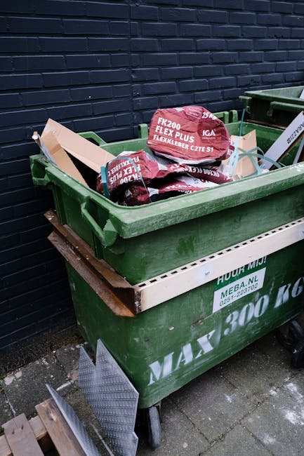 A collection of stacked cardboard fruit boxes with colourful printed labels, placed outdoors on a paved surface, alongside black plastic crates, metal wire cages filled with various cardboard boxes and packaging materials, and several closed wheelie bins in green and red. The scene appears to be part of a waste clearing operation at a residential or commercial property, with sunlight illuminating the area and a background of greenery and distant houses. The various containers and packaging suggest the site is involved in waste collection or sorting, typical of independent rubbish removal services such as those offered by House Clearance Streatham. The different textures include corrugated cardboard, smooth plastic, and metal, with the cardboard boxes showing signs of previous use and some slightly crumpled or leaning. The environment indicates an organized effort to gather materials for disposal or recycling, supporting the context of alternative waste handling methods outside local authority collection. Overall, the scene captures the process of clearing and managing bulky rubbish in preparation for safe and efficient removal, typical of private waste disposal services.