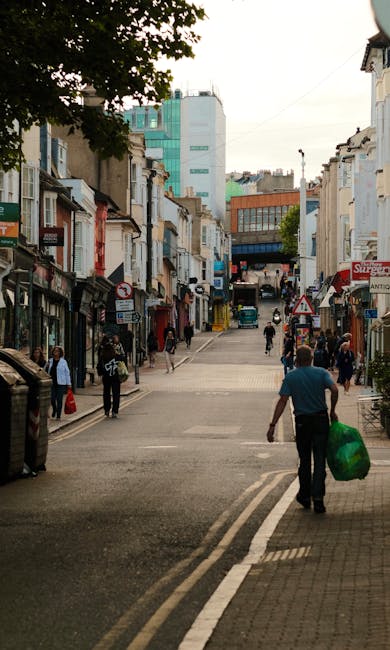 A busy urban street scene in Streatham with a gentle slope leading towards a mix of commercial and residential buildings. In the foreground, a man dressed in a blue shirt and dark trousers is walking away from the camera, carrying a green plastic bag. To his left, a parked vehicle is partially visible, and further down, pedestrians are crossing or walking along the sidewalk, some carrying shopping bags or engaged in conversation. The street is lined with various small shops, cafes, and local businesses, with shopfronts displaying signage and awnings. On the right side, a building with a pale façade and adjacent outdoor seating area can be seen. Tall modern buildings in shades of turquoise and orange are visible further in the background, contrasting with the older, shorter structures closer to street level. The scene is illuminated by natural daylight, with a large tree casting shadows over part of the street, suggesting a partly cloudy sky. The setting depicts an active locality where independent street-level access impacts the logistics of rubbish collection and waste disposal, hinting at the relevance of private rubbish clearance services such as those offered by House Clearance Streatham. The overall environment supports the context of an area where on-site or alternative waste handling may be involved, especially for busy commercial and residential neighborhoods.