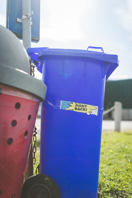 A tall, rectangular blue plastic rubbish bin with a closed lid, positioned on a paved outdoor surface next to a red rubbish container with black ventilation holes and a partially visible aluminum lid. The blue bin features a yellow and blue sticker with the words 'Fight Back!' applied slightly askew. The background includes a metal pole, possibly part of a sign or street fixture, and a blur of greenery, pavement, and a cloudy sky, indicating a street or public area environment. The scene suggests a setting for household or street waste collection, with the blue bin likely used for general waste or recycling, indicative of alternative waste handling or private rubbish removal services. House Clearance Streatham's presence in the context reinforces professional rubbish management in a residential or urban environment, with no visible action occurring in the scene.