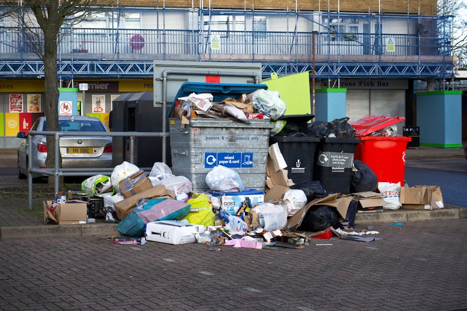 A pile of mixed household rubbish spread across a flat surface, consisting of paper, cardboard, plastic bottles, and crumpled packaging, with some bottles featuring labels and caps. The paper and cardboard are creased and torn, while the plastic bottles vary in size and shape, with some lying on their sides. The rubbish appears to include both recyclable materials and general waste, with some items partially overlapping or stacked. The background is blurred, suggesting an indoor or outdoor environment designated for waste collection or disposal, consistent with the context of private rubbish removal services offered by House Clearance Streatham. The lighting is even, highlighting the textures of the different materials, from smooth plastic to rough paper, giving a clear view of typical household waste accumulated in preparation for removal or recycling. This scene visually supports the process of professional rubbish collection, emphasizing the natural assortment of waste materials handled during house or site clearance activities.